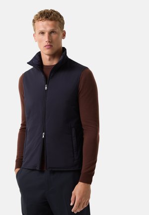 Gilet sans manches bleu marine avec col montant, fermeture éclair à l'avant et poches latérales. Fabriqué à partir d'un tissu lisse et léger. Porté par-dessus un pull sombre.