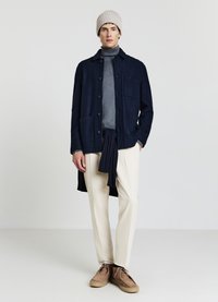 Giacca in lana blu navy con chiusura a bottoni e tasche frontali, dolcevita grigio, pantaloni crema, scarpe marroni e un berretto beige lavorato a maglia.