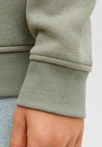 Žalias medvilnės džemperio rankogalis su raukšlėtu tekstūru. Po rankogaliu matoma ranka, rodanti odos atspalvį. Siūlės detalė rankogalyje. Kasdienis dizainas.