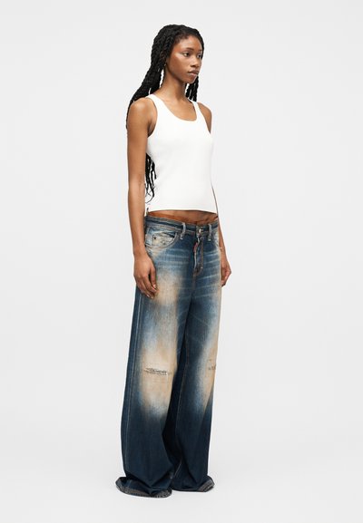 Νεαρή γυναίκα με μακριά κοτσίδα, φοράει λευκό αμάνικο τοπ και φαρδιά, ξεθωριασμένα μπλε τζιν με κοντερεμένες φθορές, στέκεται πλάγια.