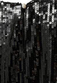 Sort paliet beklædning med firkantede paletter, der skaber en struktureret overflade, og indeholder en lille hvid knaplukning ved halsudskæringen.