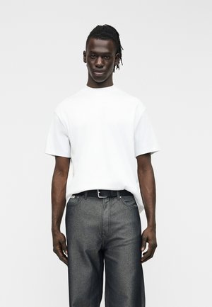 Man met donkere huid en dreadlocks draagt een eenvoudig wit kortärmelig shirt, ingestopt in een grijze high-waist spijkerbroek met een zwarte riem, staand tegen een witte achtergrond.