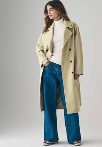Khaki värvi trench coat, millel on topelt nööbid, kombineeritud kõrge vöökohaga siniste laiade pükstega ja valgete sneakeritega. Sile, tekstureeritud kangas.