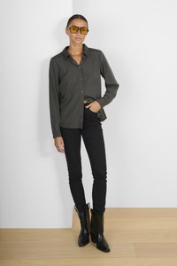 Camicia grigia a maniche lunghe con bottoni, jeans neri aderenti e stivali da cowboy neri. Grandi occhiali da sole giallo-tinti completano il look.