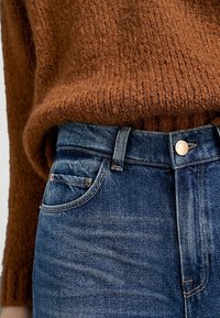Maglione a coste marrone sopra jeans di denim blu scuro, con un taglio a vita media, tasche anteriori e chiusura a bottone in ottone.