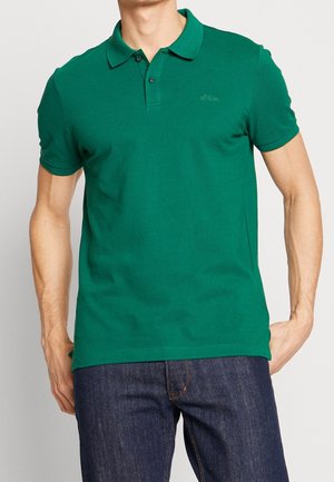 Hombre con una camiseta polo verde lisa de manga corta y vaqueros azul oscuro, de pie con una mano en la cadera contra un fondo liso.