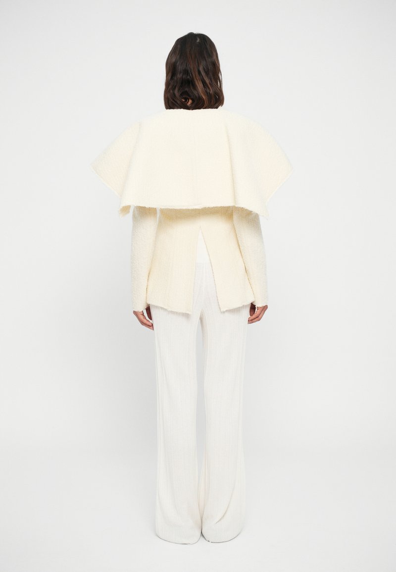 Cremfarbener Cape-Jacke mit hohem Kragen, geschichtetem Rückendesign, kombiniert mit fließenden weißen weiten Hosen.