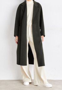 Abrigo largo verde con solapas muesca, bolsillos laterales, combinado con un jersey de canalé crema y pantalones anchos crema, llevado con zapatillas blancas.