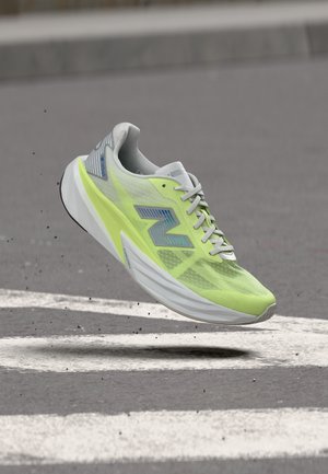 Lichtgroene en grijze New Balance hardloopschoen in de lucht boven asfalt met witte wegmarkeringen.