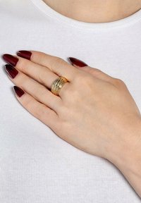 Set di anelli impilati color oro su una mano con pelle chiara, caratterizzato da più bande testurizzate. Unghie lunghe e appuntite di colore rosso scuro. Sfondo bianco della camicia.
