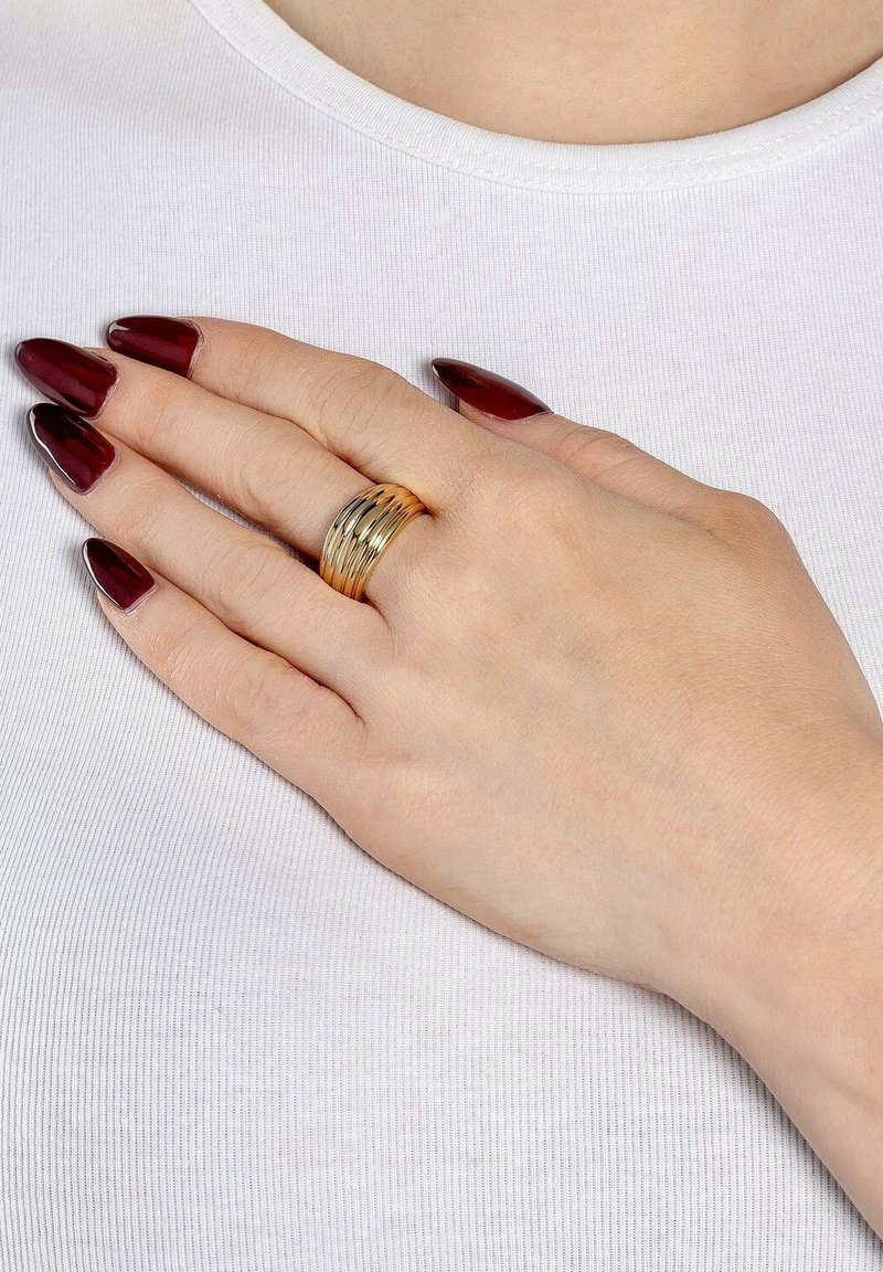 Set di anelli impilati color oro su una mano con pelle chiara, caratterizzato da più bande testurizzate. Unghie lunghe e appuntite di colore rosso scuro. Sfondo bianco della camicia.