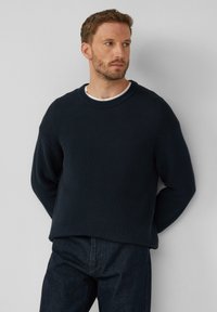 Mornarsko modra pletena pulover s okroglim izrezom in spuščeno rameni, kombiniran z temnimi jeansi. Tekstura se zdi mehka in debela.