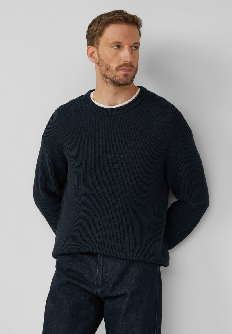 Mornarsko modra pletena pulover s okroglim izrezom in spuščeno rameni, kombiniran z temnimi jeansi. Tekstura se zdi mehka in debela.