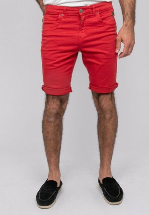 Homme portant un short rouge vif remonté et des chaussures noires sans lacets, debout devant un fond uni.