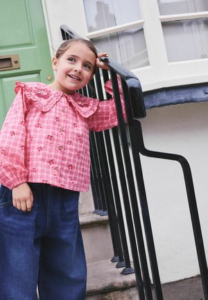 Młoda dziewczyna w różowej bluzce z falbankowanym kołnierzem opiera się o czarną balustradę przy zewnętrznych schodach i zielonych drzwiach, uśmiecha się i patrzy w górę.