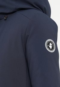 Námořnická modrá nepromokavá bunda s hladkým povrchem, která má na rukávu nášivku s logem a nastavitelnou kapuci s šňůrkovými detaily.