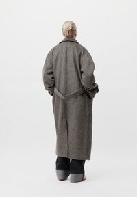 Άτομο με ανοιχτά μαλλιά σε κότσο, φορώντας ένα μακρύ καρό παλτό, φαρδιά παντελόνια με μανίκια και λευκά παπούτσια, σε φόντο λευκό.