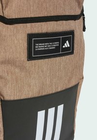 Brauner Stoffrucksack mit einem schwarzen Bodenpanel und einem auffälligen Logo-Plakette mit drei weißen Streifen und Text in mehreren Sprachen.