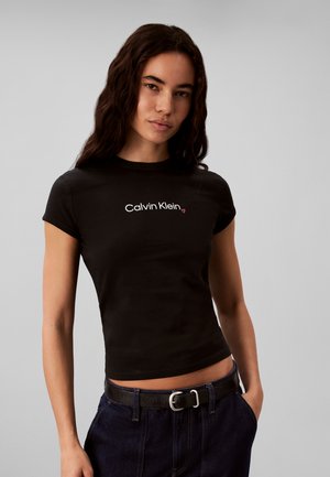 Kobieta z długimi ciemnymi włosami, ubrana w dopasowany czarny t-shirt Calvin Klein i ciemne jeansy z czarnym paskiem, stojąca z jedną ręką w kieszeni.