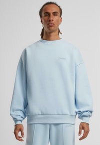 Helles blaues, übergroßes Sweatshirt aus einem weichen Baumwollmix. Verfügt über einen Rundhalsausschnitt, überschnittene Schultern und ein dezentes, gesticktes Logo auf der Brust.