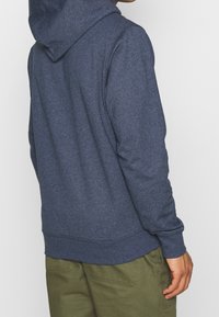 Mörkblå hoodie i mjukt material, med snörhätta, långa ärmar och ribbade muddar och avslut. Bärs med olivgröna byxor.