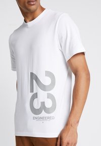 Camiseta de algodón blanca con gráficos en gris bold de "23" y "ENGINEERED" en la parte delantera, con cuello redondo y mangas cortas.