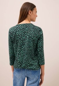 Femme aux longs cheveux bruns portant un haut à motifs noir et vert à manches longues et un jean bleu, vue de dos sur un fond uni.