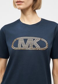 Γυναίκα που φοράει μπλε ναυτικό T-shirt με χρυσό διακοσμητικό λογότυπο "MK" στο κέντρο του στήθους, κοντά μανίκια και στρογγυλή λαιμόκοψη.