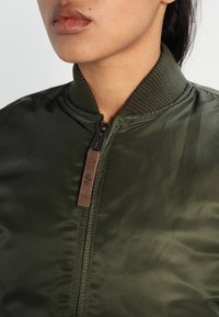 Oliwkowa kurtka bomberka z prążkowanym kołnierzem, zamkiem błyskawicznym z przodu i skórzaną zawieszką. Gładki, błyszczący materiał z subtelnymi detalami tekstury.