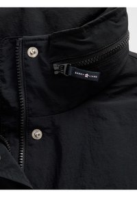 Gros plan sur le col d'une veste noire avec une fermeture éclair et des boutons-pression, montrant une languette de tirette bleu marine portant l'inscription "Sandy Liang" et un petit logo "Gap".