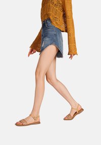 Sandali beige con cinturini intrecciati, suola piatta e fibbia alla caviglia. Abbinati a una maglietta gialla con texture e una gonna di jeans, che mette in mostra un look casual.