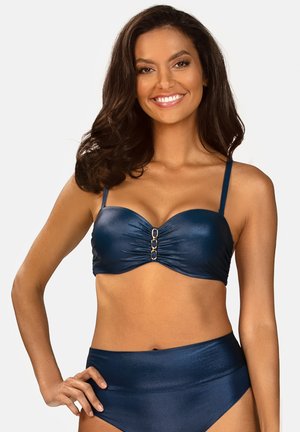 Uśmiechnięta kobieta z długimi ciemnymi włosami, ubrana w granatowy bikini z ozdobnymi złotymi pierścieniami, pozująca z jedną ręką na biodrze.