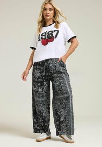 Witte t-shirt met zwarte rand en een graphic "1987", gecombineerd met wijde zwarte broek met witte paisleypatronen en een trekkoord in de taille.