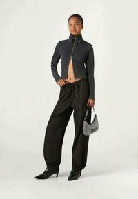 Juodas trumpas švarkas su aukštu koloriumi ir priekine užtrauktuku, derinamas su laisvais juodais kelnėmis ir tekstūruota pilka rankine su grandinės dirželiu.