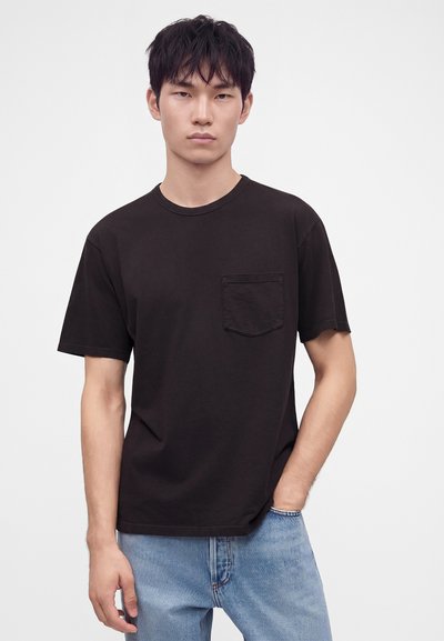 Jeune homme aux cheveux courts et foncés, portant un t-shirt noir avec poche et un jean bleu clair, debout avec une main dans la poche.