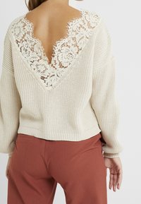 Frau in einem cremefarbenen, gestrickten Pullover mit tiefem V-Rücken und Spitzen-Detail, getragen mit rostfarbenen Hosen, von hinten gezeigt.