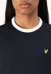 T-shirt bleu marine avec un col côtelé blanc et une petite broderie d'aigle jaune sur la poitrine. Texture lisse, coupe régulière.