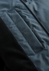 Close-up of a dark jacket showing a vertical zipper, a padded fabric panel, and a textured fleece section.