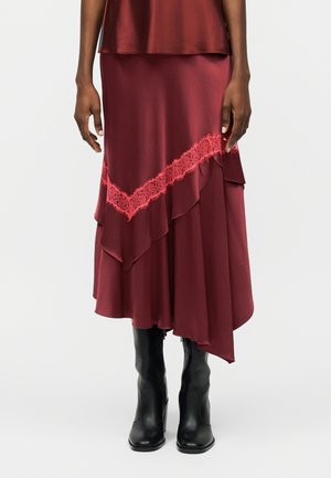 Μοντέλο φοράει μπορντό ασύμμετρη φούστα με στρώσεις βολάν και κόκκινη δαντέλα στη φάσα, συνδυασμένη με μαύρα δερμάτινα μποτάκια μέχρι τη μέση της γάμπας.