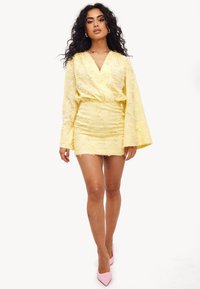 Robe enveloppante jaune avec un motif floral texturé, manches longues en forme de cloche et un ourlet court. Portée avec des escarpins roses à bout pointu.