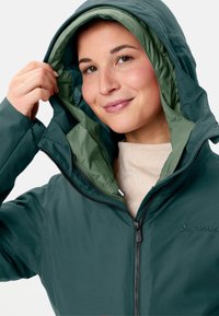 Giacca isolante verde scuro con cappuccio a doppio strato, zip frontale e logo. Esterno liscio con fodera interna morbida di un verde più chiaro.