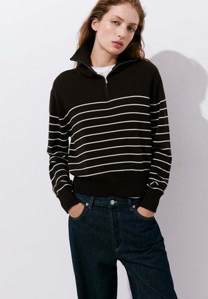 Mujer joven con las manos en los bolsillos lleva un suéter negro con rayas horizontales blancas y vaqueros azul oscuro sobre un fondo liso y claro.