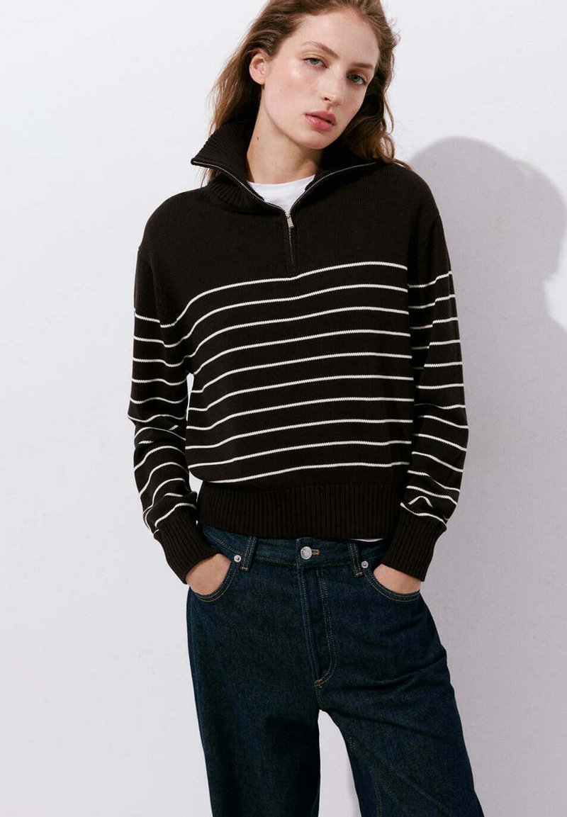 Jeune femme les mains dans les poches porte un pull noir à rayures blanches horizontales et un jean bleu foncé sur un fond clair uni.