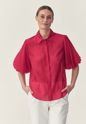 Kobieta o jasnobrązowych włosach, ubrana w czerwoną bluzkę z krótkimi bufianymi rękawami i białe spodnie, stojąca z rękami w kieszeniach.