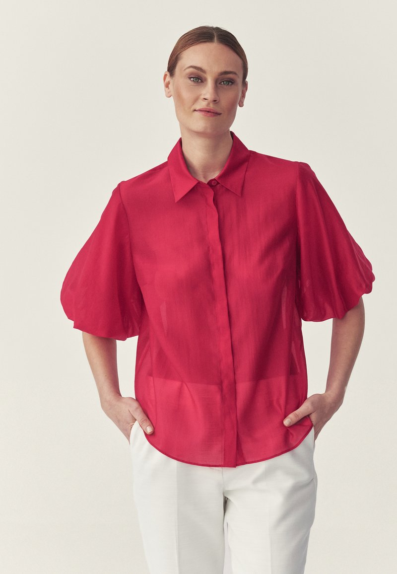Femme aux cheveux châtain clair portant un chemisier rouge à manches courtes bouffantes et un pantalon blanc, debout les mains dans les poches.