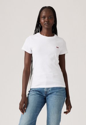 T-shirt en coton blanc à manches courtes, encolure ronde et petit logo rouge sur la poitrine gauche ; porté avec un jean en denim bleu.