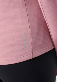 Nahaufnahme des Saums eines rosa Langarm-Tops mit vertikalem "Swedemount"-Logo und sichtbaren Nähten, kombiniert mit einer schwarzen Hose.
