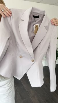 Vaaleanharmaa muotopukine, jossa on kaksi koristeellista kultanappia, pidetään käsissä, joissa on sormuksia. Taustalla on sisätilat, joissa on puulattia ja kasveja.