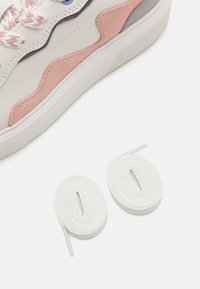 White leather sneaker with pink suede accents, textured cream sole, and colorful braided laces. Two additional plain white lace pairs included.