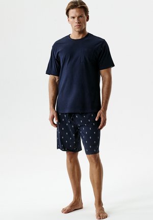 Mężczyzna w granatowej koszulce z krótkim rękawem i wzorzystych granatowych szortach, stojący boso na jednolitym jasnym tle.